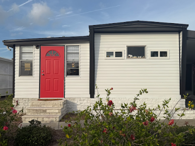 White house with red door, small steps, bushes, and a blue sky.