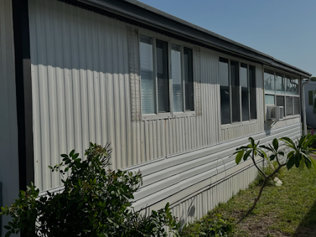 White corrugated siding building with multiple windows, grass, and plants.