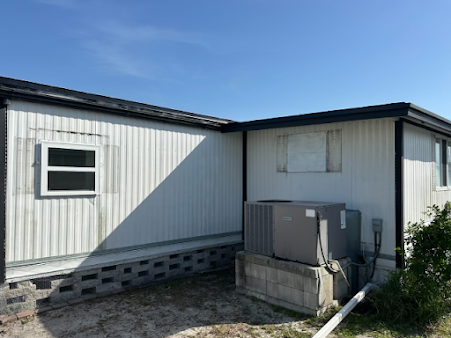 Exterior of a weathered white modular building with dark trim, an air conditioning unit, and a blue sky.