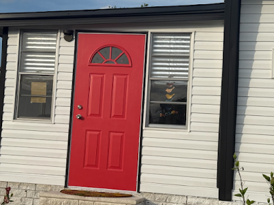 Red door with a half-circle window flanked by two windows with blinds on a white house with black trim.