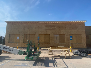Building under construction with plywood walls, blue sky, parking lot, and construction equipment.