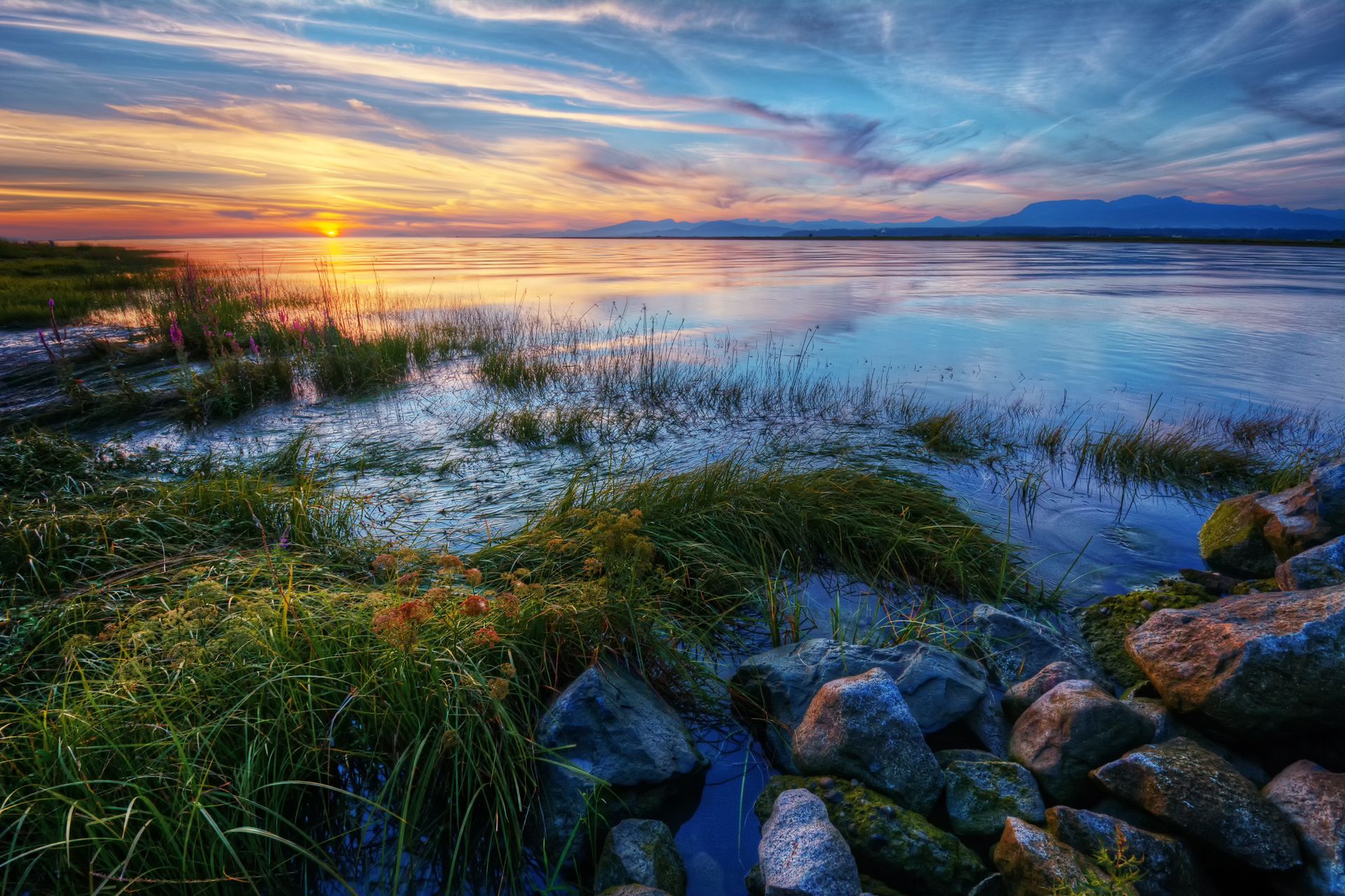 A sunset over a lake with rocks in the foreground and mountains in the background.