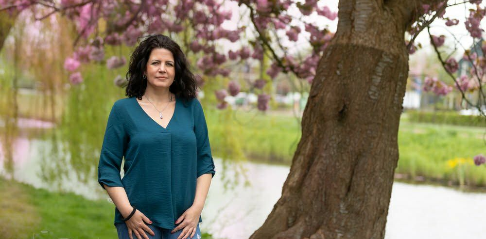 Vrouw in een turquoise top staat naast een boom bij een vijver met roze bloesem op de achtergrond.