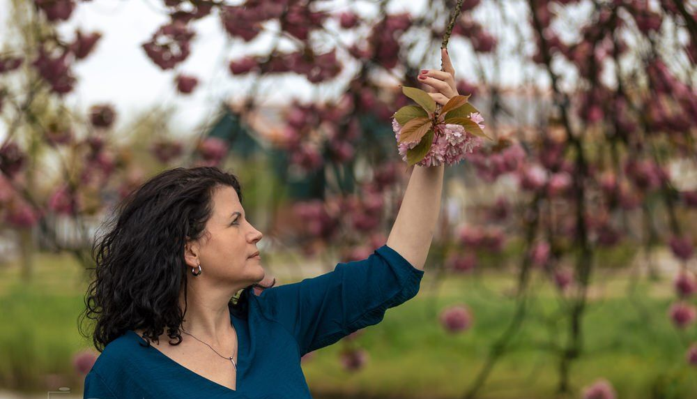 Vrouw in een blauwe top reikt omhoog om roze bloesems in een tuin aan te raken.