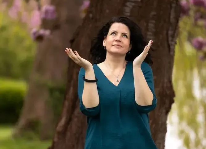 Vrouw in een turquoise shirt met opgeheven handen, staand buiten bij een boom met roze bloesem.