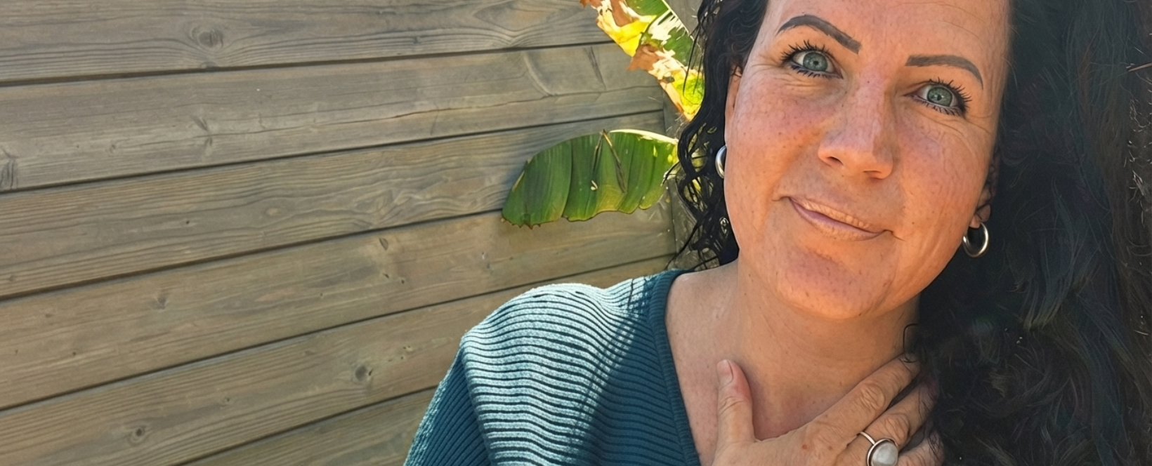 Vrouw in een turquoise trui, lachend buiten naast een houten muur in het zonlicht.