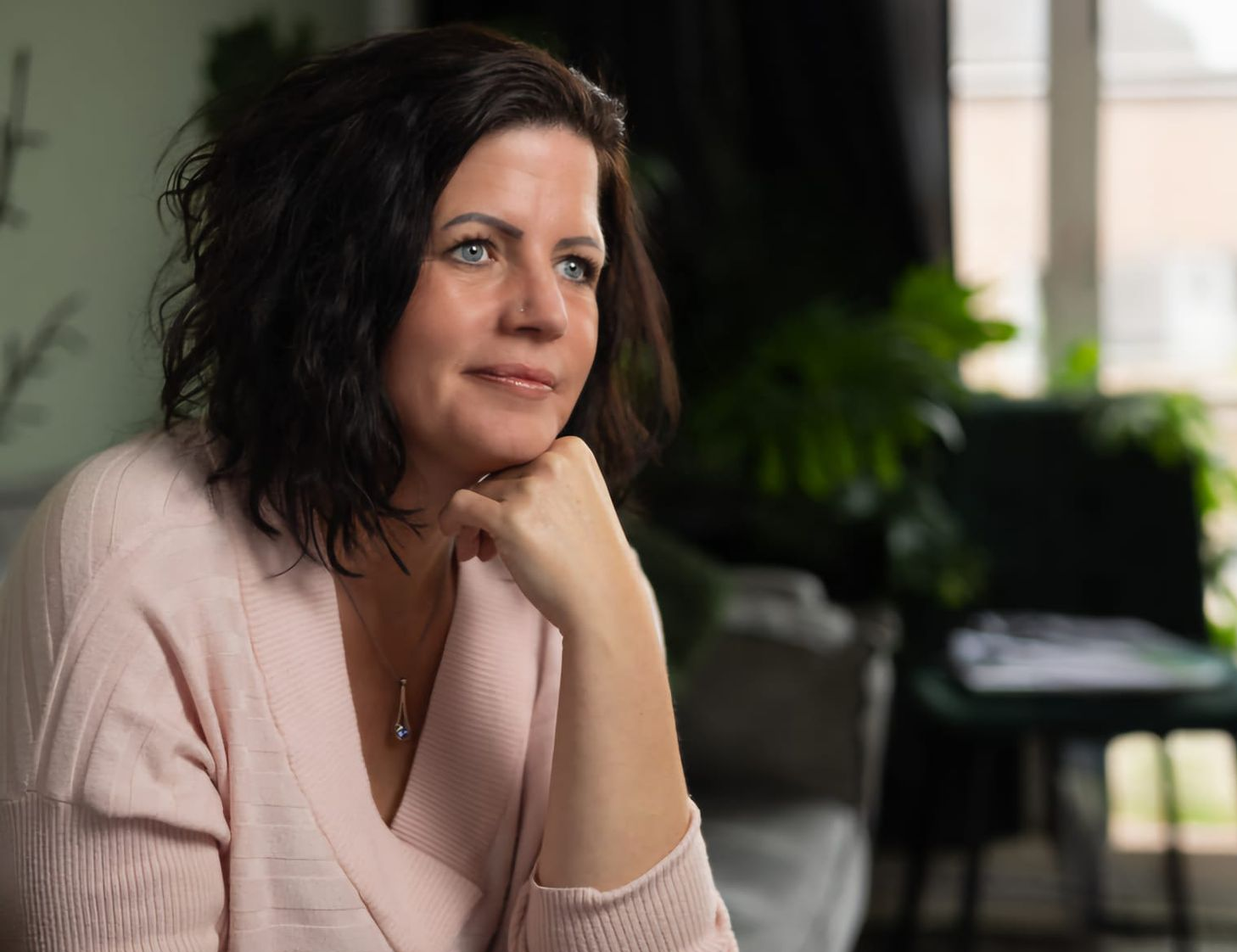 Een vrouw in een roze trui rust met haar kin op haar hand en staart peinzend in een zacht verlichte kamer.