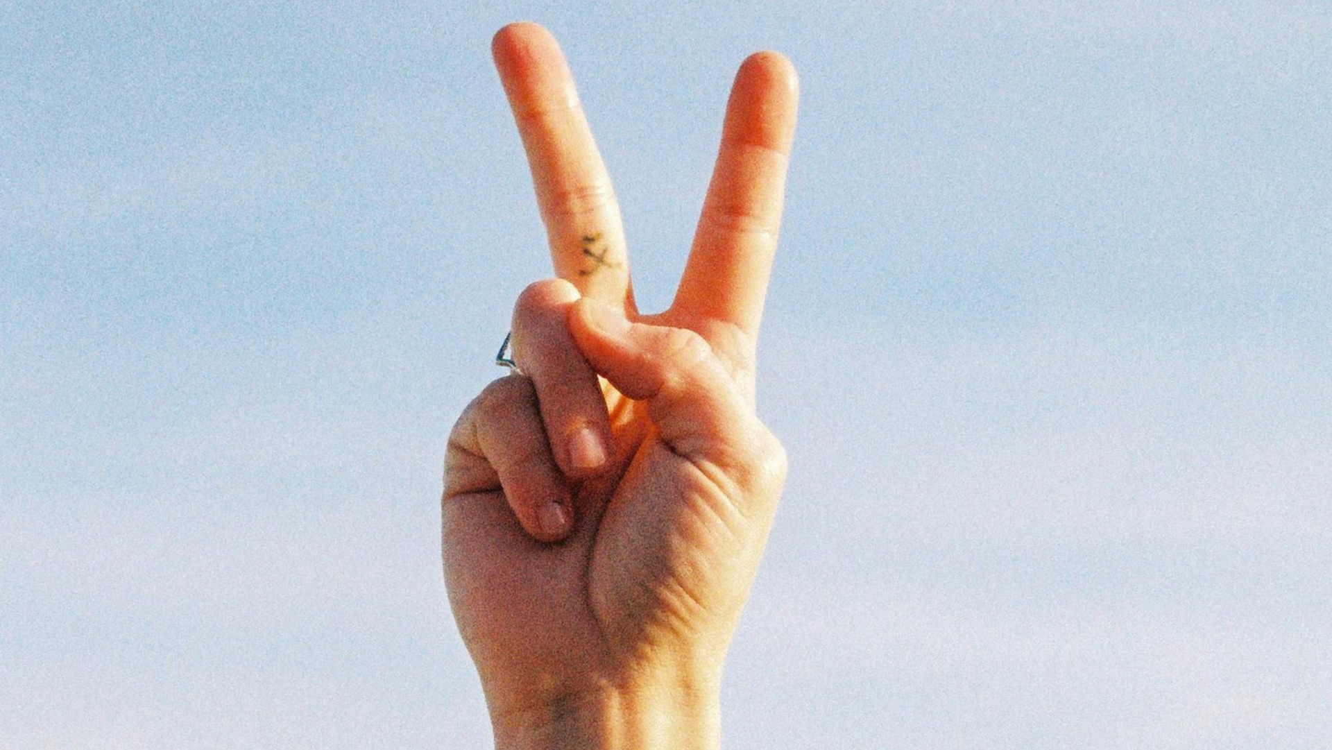 Een opgeheven hand die een vredesteken maakt tegen een blauwe lucht, met een kleine tatoeage op de pols.