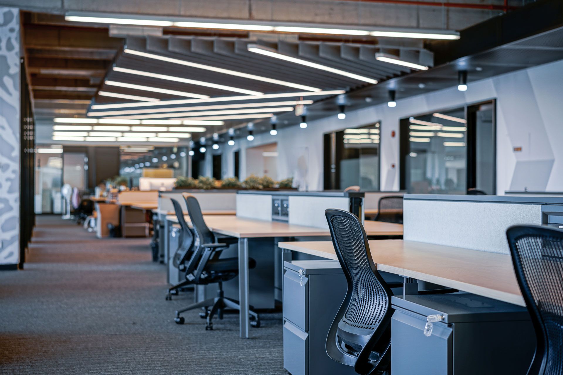 Modern open-plan office with desks, swivel chairs, and pendant lighting, viewed down a long workspace corridor