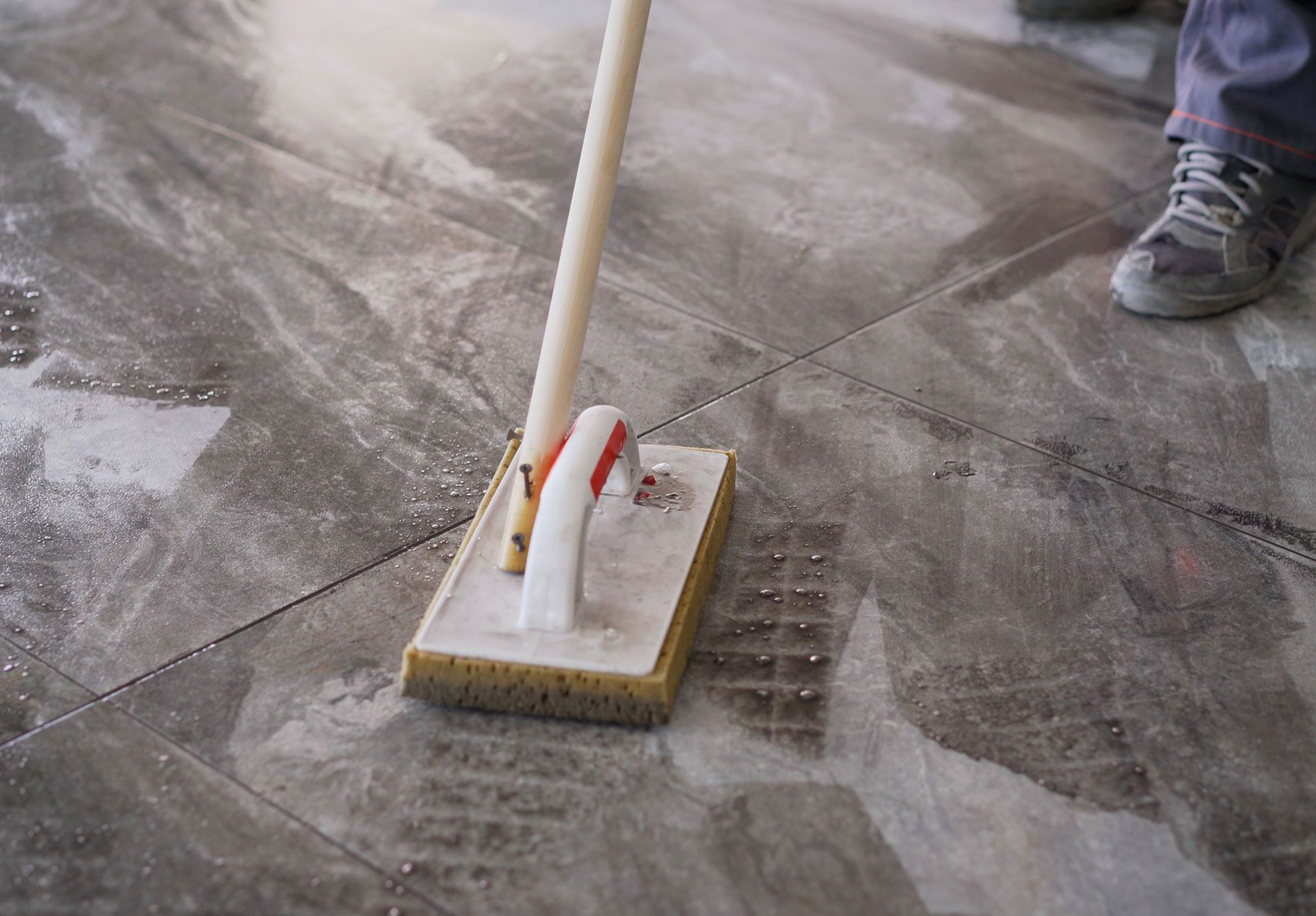 Scrubbing a concrete floor with a brush on a wet surface, with a person’s shoe visible nearby