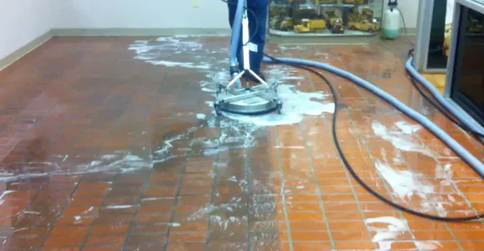 Floor cleaning machine scrubbing a tiled floor, leaving white foam behind, with a hose trailing across it.