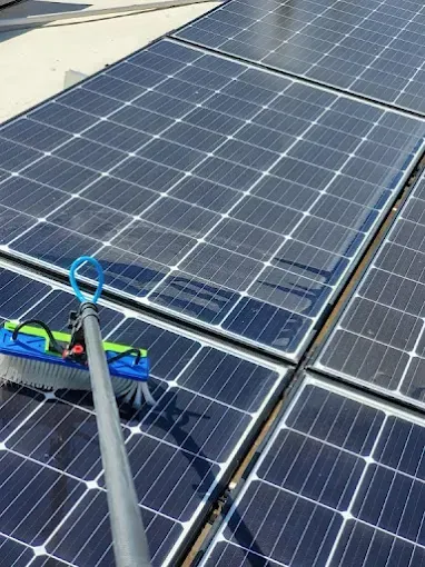 Person cleaning rooftop solar panels with a long-handled brush under a blue sky