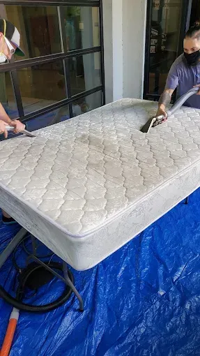 Two people cleaning a mattress outdoors on a blue tarp with a hose or pressure washer
