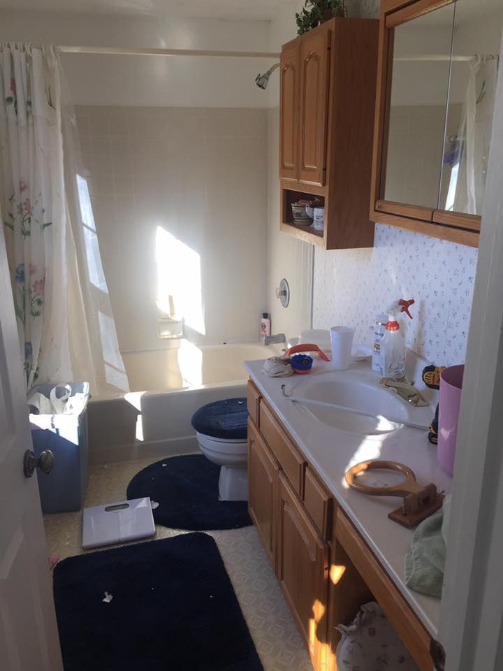 A small bathroom with light wood cabinets, a white sink, a toilet with a dark mat, and a bathtub with a floral curtain.