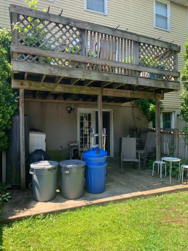A wooden deck attached to the back of a house, above a patio area with trash bins and outdoor furniture.