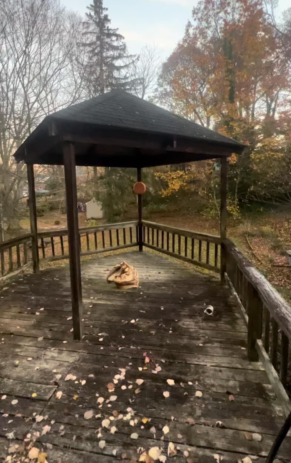 An outdoor wooden gazebo with a dark roof stands on a weathered deck covered in scattered autumn leaves.