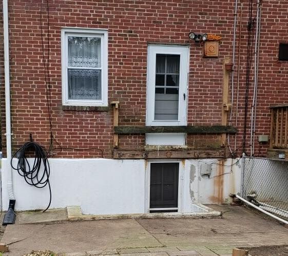A brick exterior wall with a white-framed window, a white door mounted above a basement door, and a coiled hose on the left.