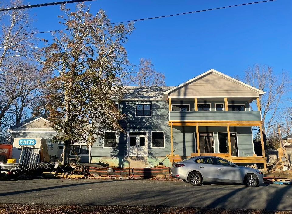 A two-story blue house under construction with an unfinished wooden porch, parked car in front, and trees against blue sky.