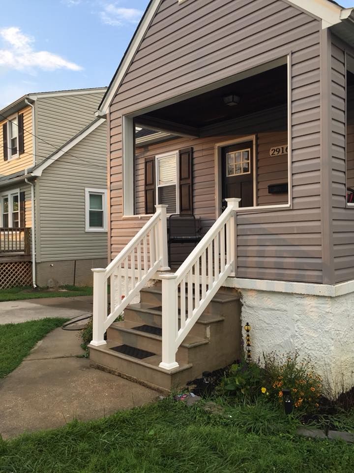 Gray house with a porch, white railing, and stairs leading to the front door, next to a beige house on a grassy street.