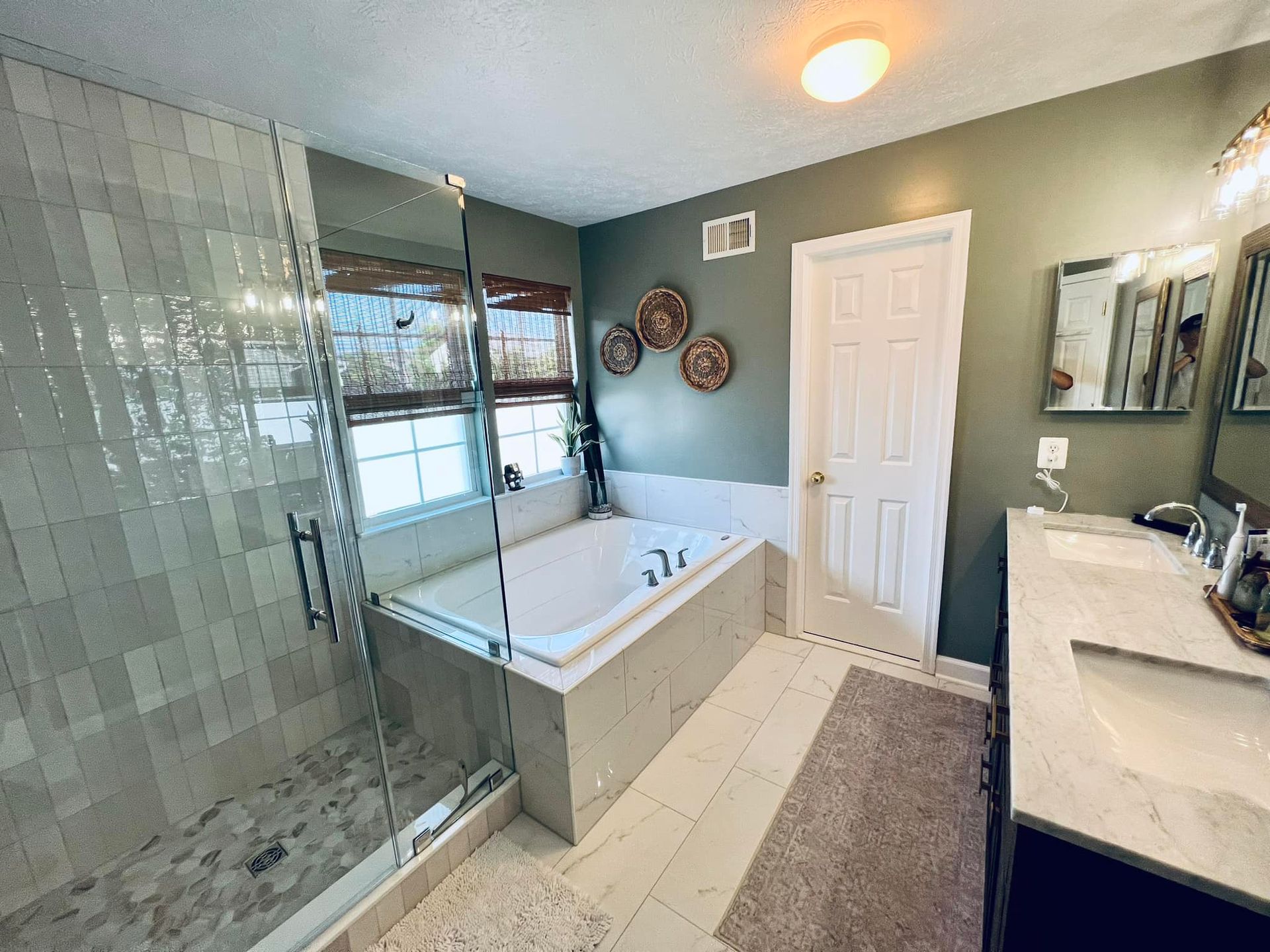 A modern bathroom with a glass shower, a drop-in bathtub against a sage green wall, and a double vanity.