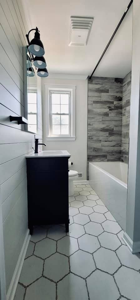 A small, modern bathroom with white shiplap walls, a black vanity, hexagon floor tiles, and a gray stone-tiled bathtub.