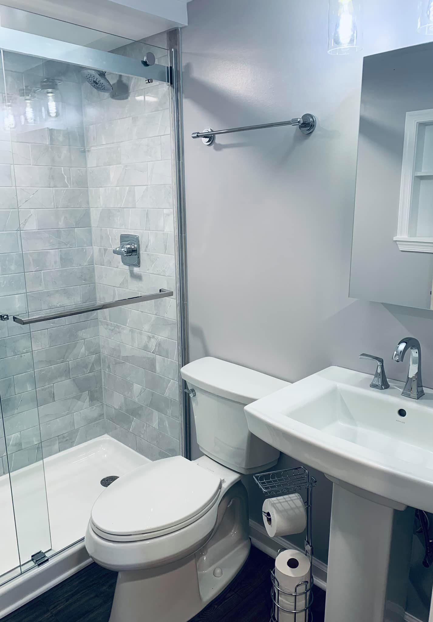 A modern bathroom with a white pedestal sink, toilet, and a glass-enclosed shower with tiled walls and light gray paint.