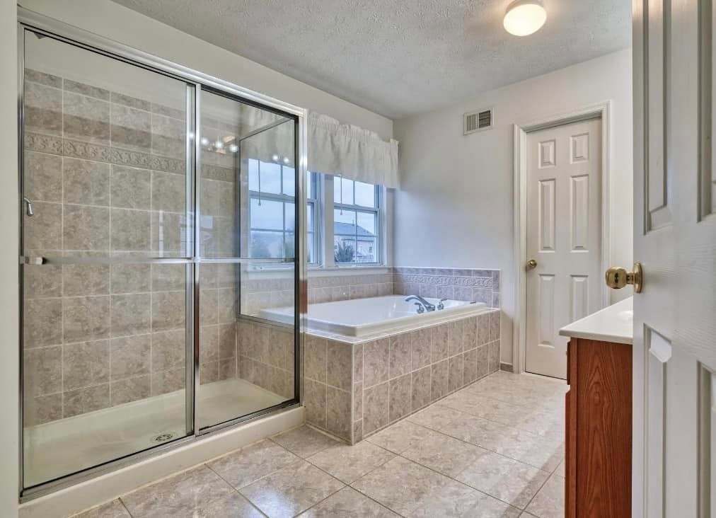 A beige tiled bathroom with a glass-enclosed shower, a built-in soaking tub, a window, and a door.