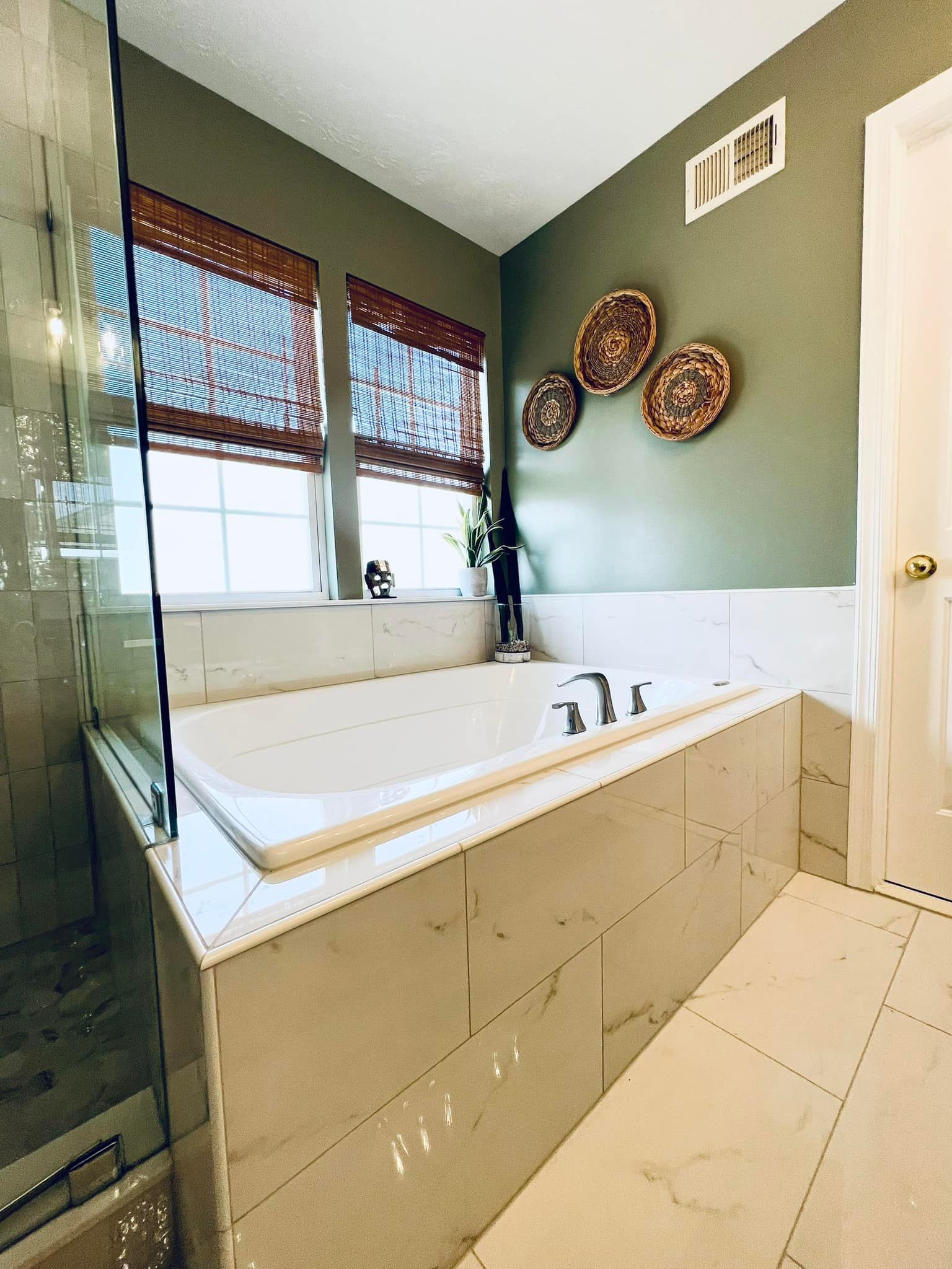 A soaking tub in a bathroom with sage green walls, tiled surround, and three decorative woven wall baskets.