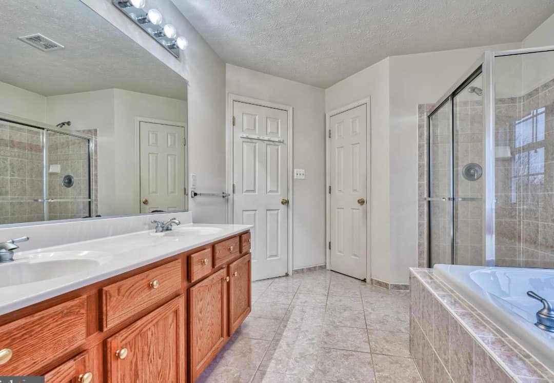 A brightly lit bathroom with a double wood vanity, large mirror, separate shower, and a built-in bathtub on tiled floors.