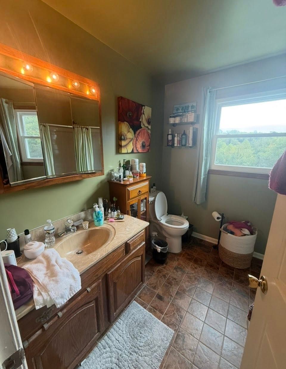 A bathroom with green walls, a wooden vanity, a beige sink, a white toilet, tiled flooring, and a window with a view.