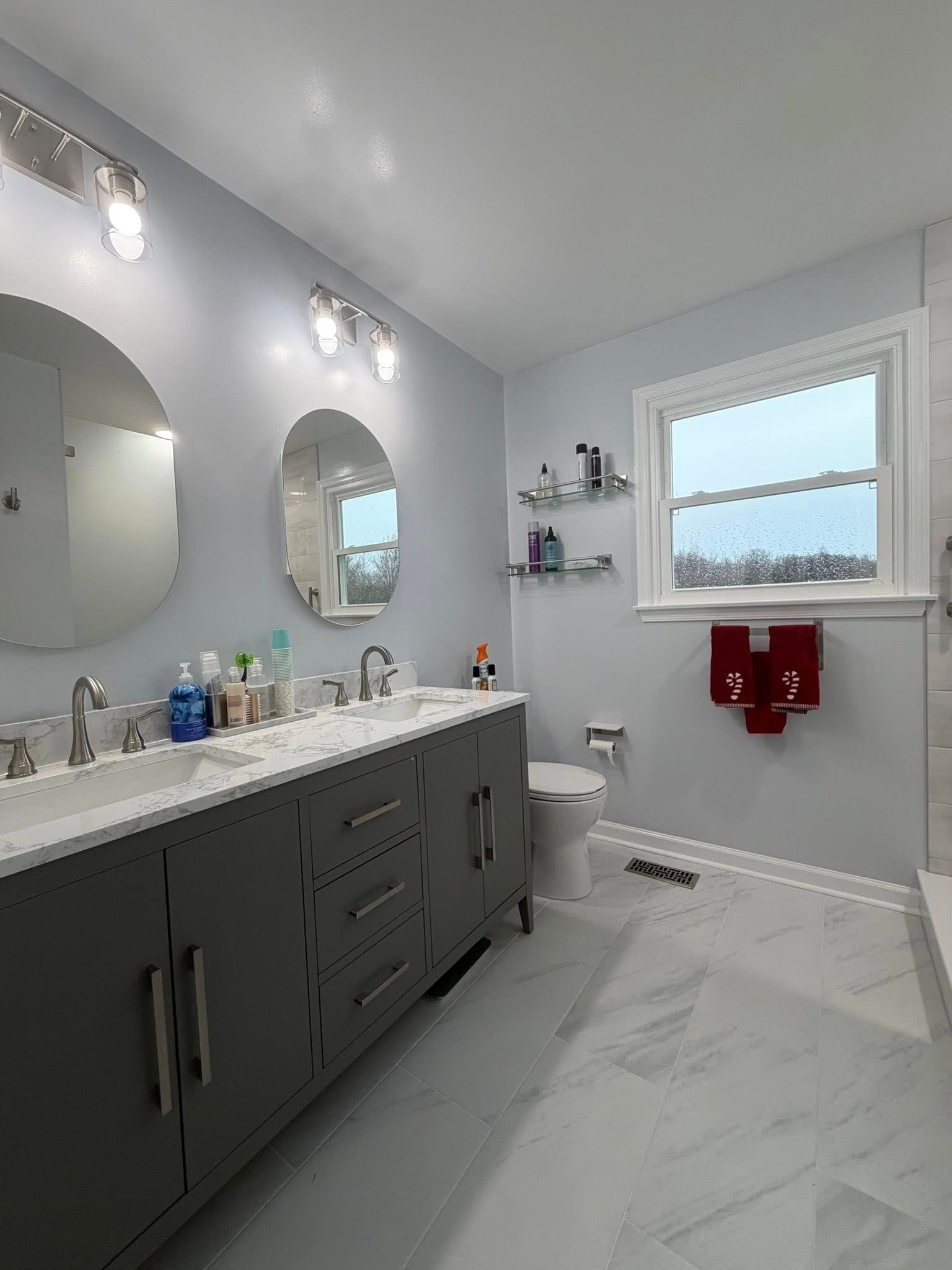 A modern bathroom with a dark gray dual-sink vanity, two oval mirrors, white marble tile flooring, and a white toilet.