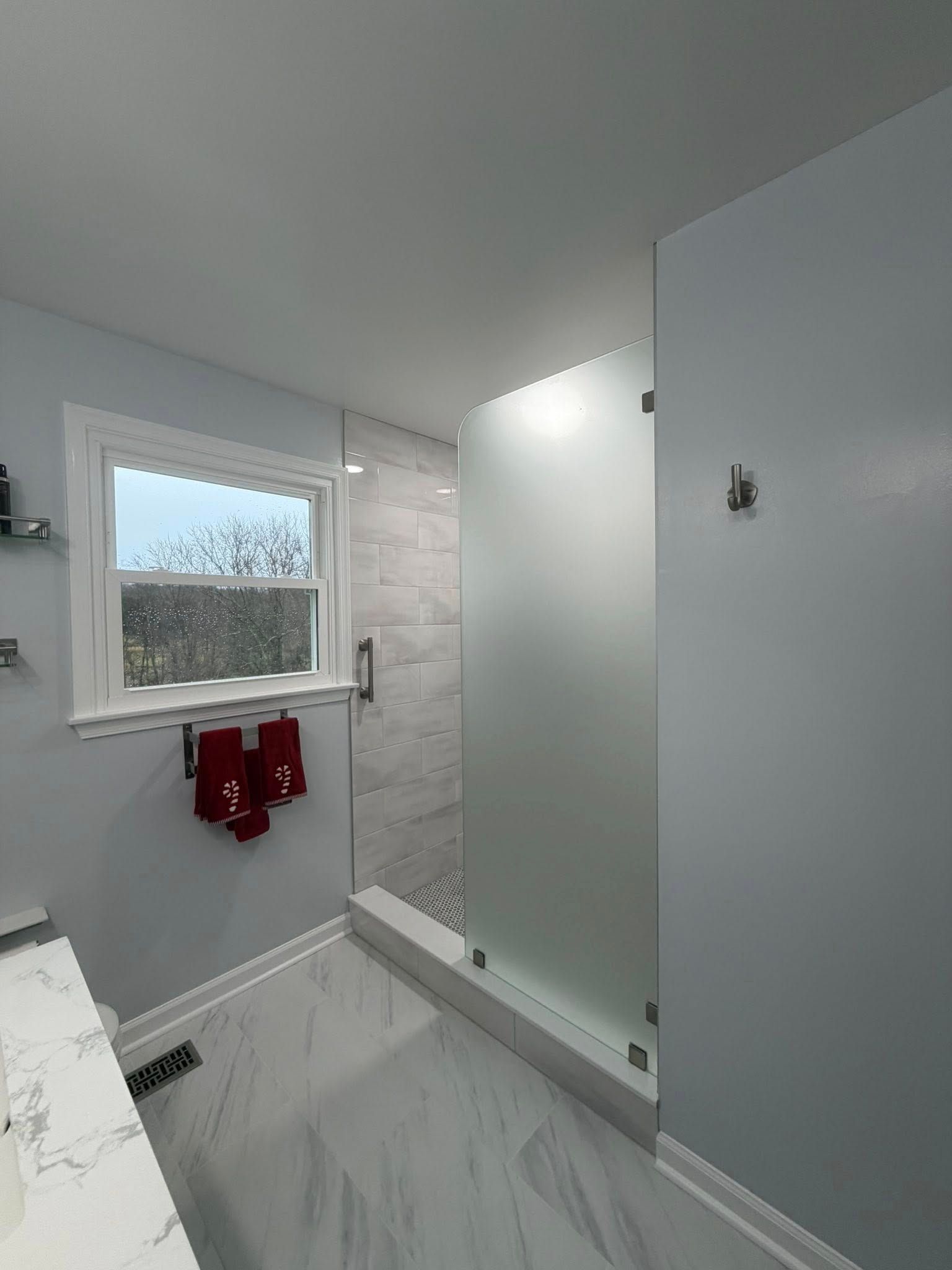 A bathroom with a frosted glass shower, marble-patterned tile floor, light blue walls, and a window with red towels below it.