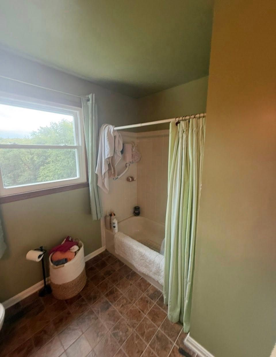A bathroom with sage green walls, a window, a white tub with a light green shower curtain, and a laundry basket on the floor.
