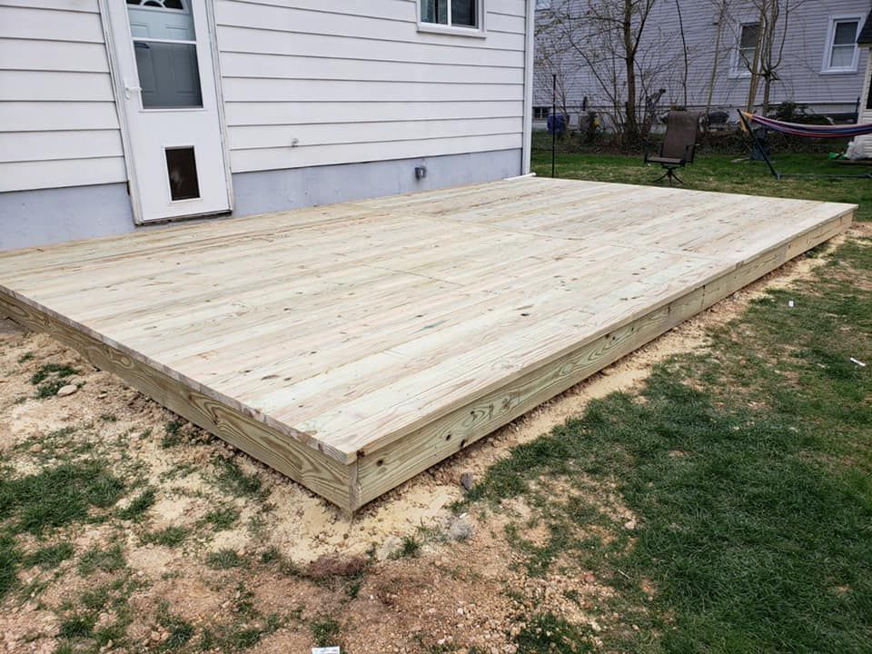 A newly constructed wooden deck attached to the side of a white house, situated on a yard with dirt and grass.