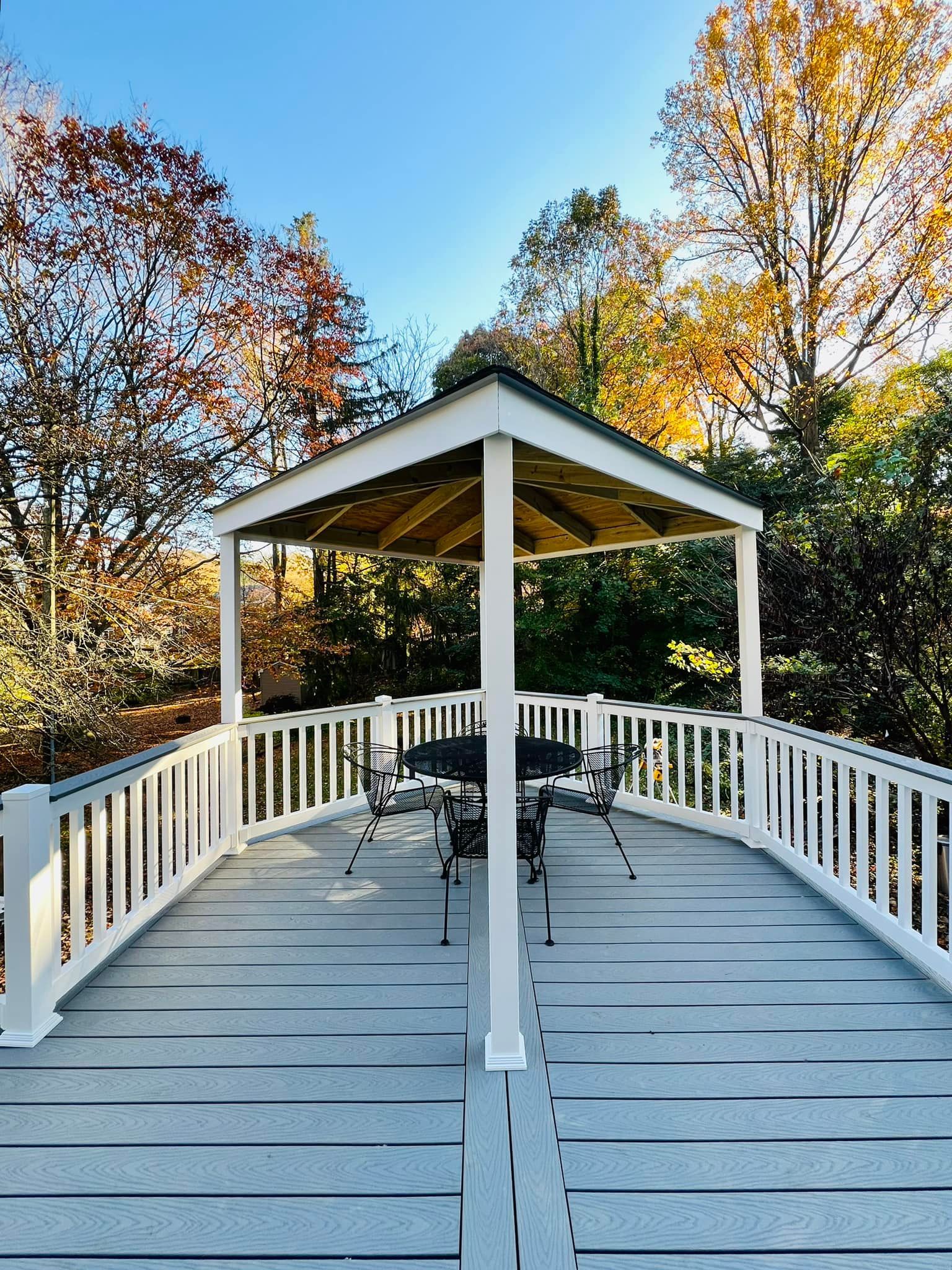 A white pergola over a patio set on a gray deck, surrounded by autumn trees under a clear blue sky.