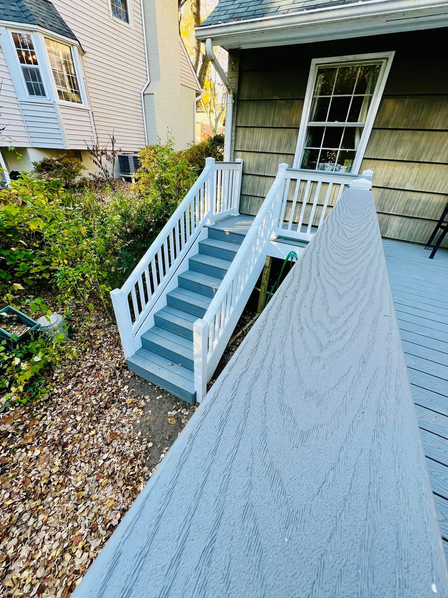 A grey deck with white railings leads to a set of matching grey stairs descending to the ground beside a house.