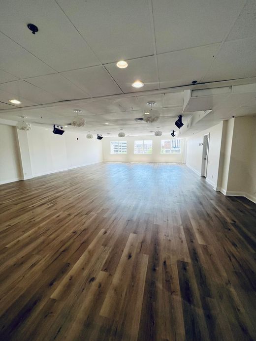 A large, empty studio with light wood floors, white walls, recessed ceiling lights, and windows at the far end.