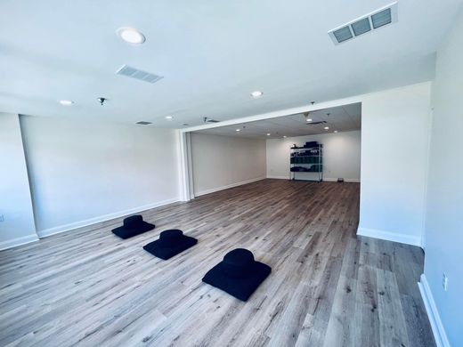 A wide, minimalist yoga studio with wood-look floors, white walls, and three black meditation cushions on the floor.
