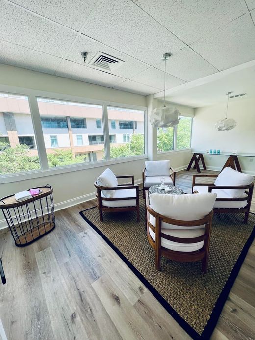 A bright seating area with four white chairs arranged around a round rug on light wood floors, next to a long window.