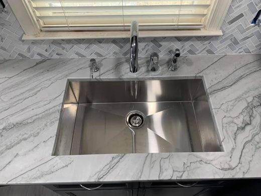 A stainless steel kitchen sink with a chrome faucet set in a white, marble-patterned countertop against a tiled backsplash.
