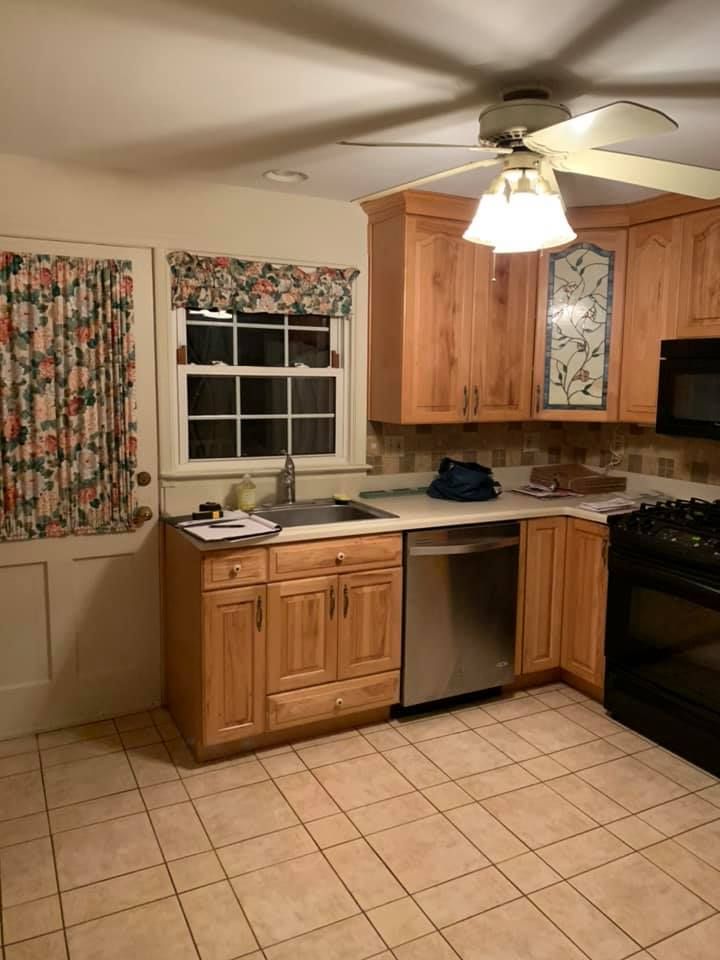 A kitchen with light wood cabinets, stainless steel dishwasher, tiled floor, and floral curtains on a window and door.