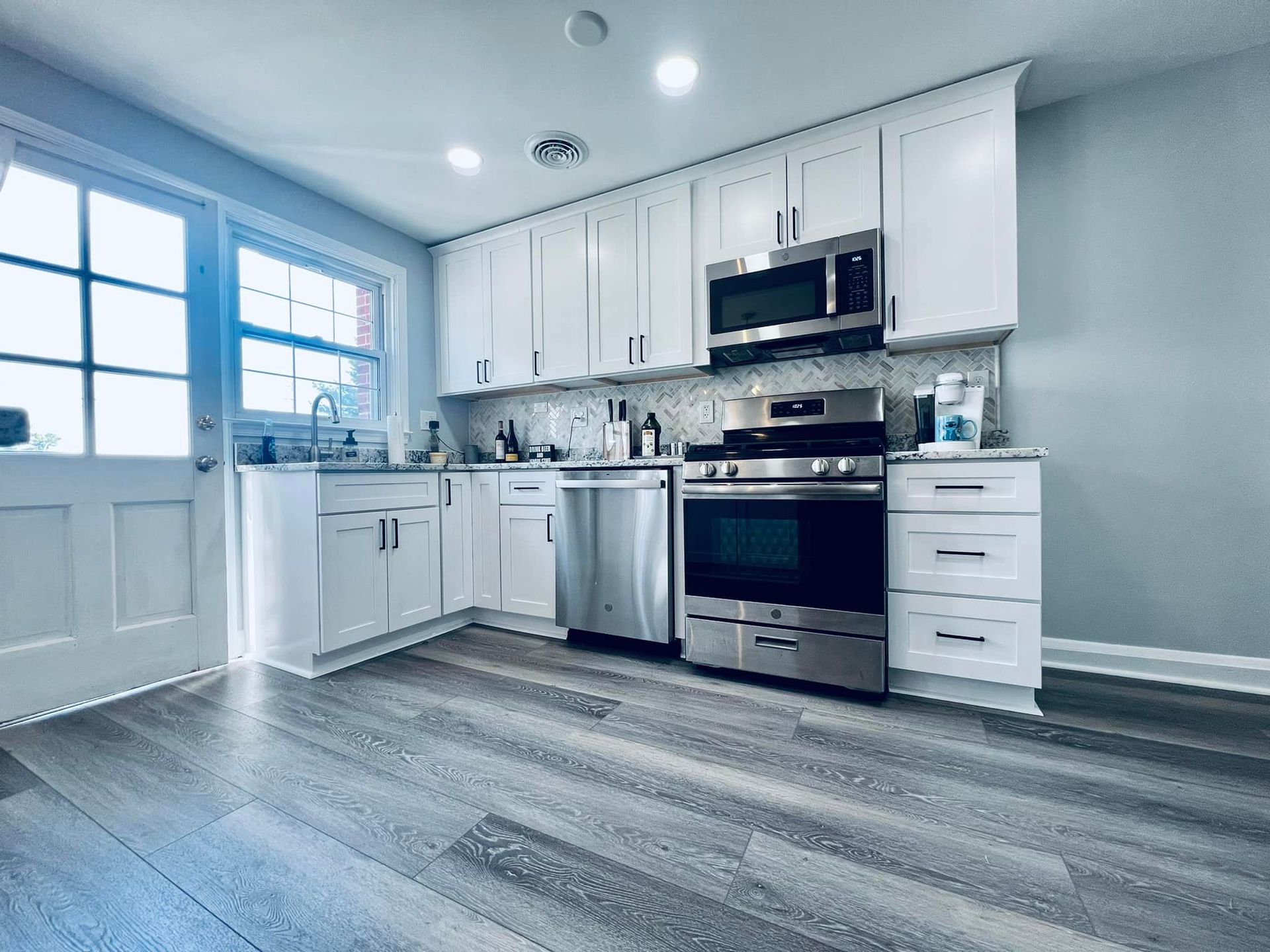 A modern kitchen with white cabinets, stainless steel appliances, and grey wood-look flooring near a glass-paned door.