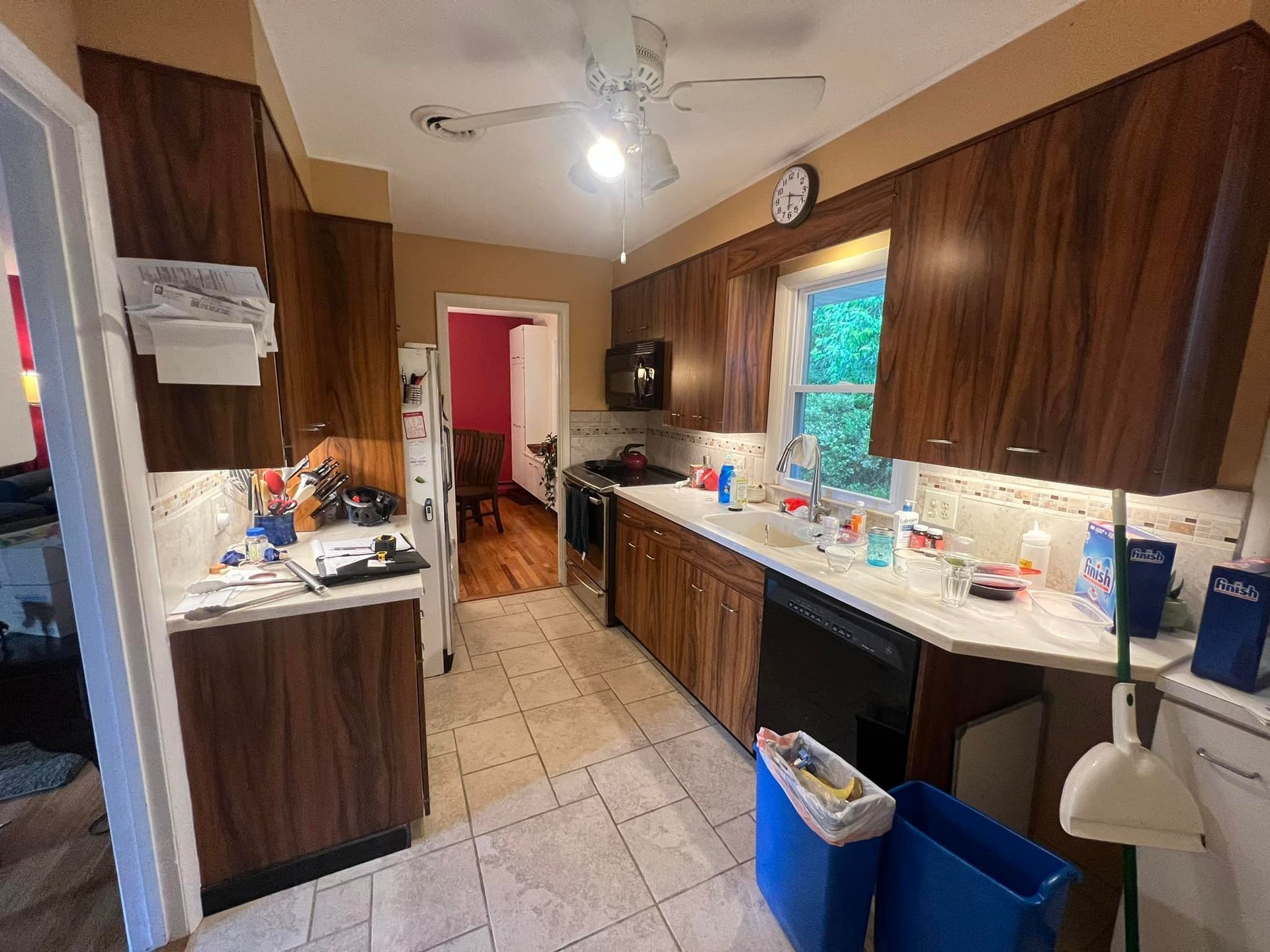 A kitchen with wood cabinets, light-colored countertops, tiled flooring, a ceiling fan, and a view into a dining room.