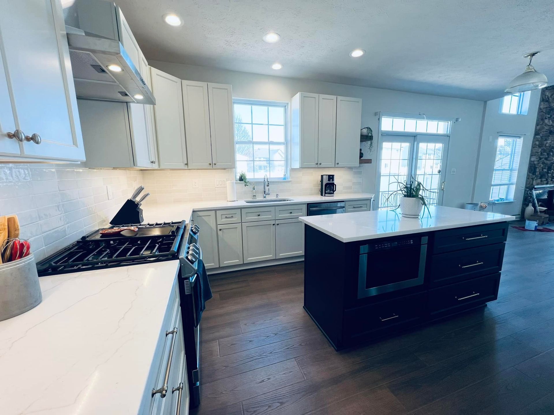 A modern, brightly lit kitchen with white cabinetry, white countertops, a dark blue center island, and wood floors.