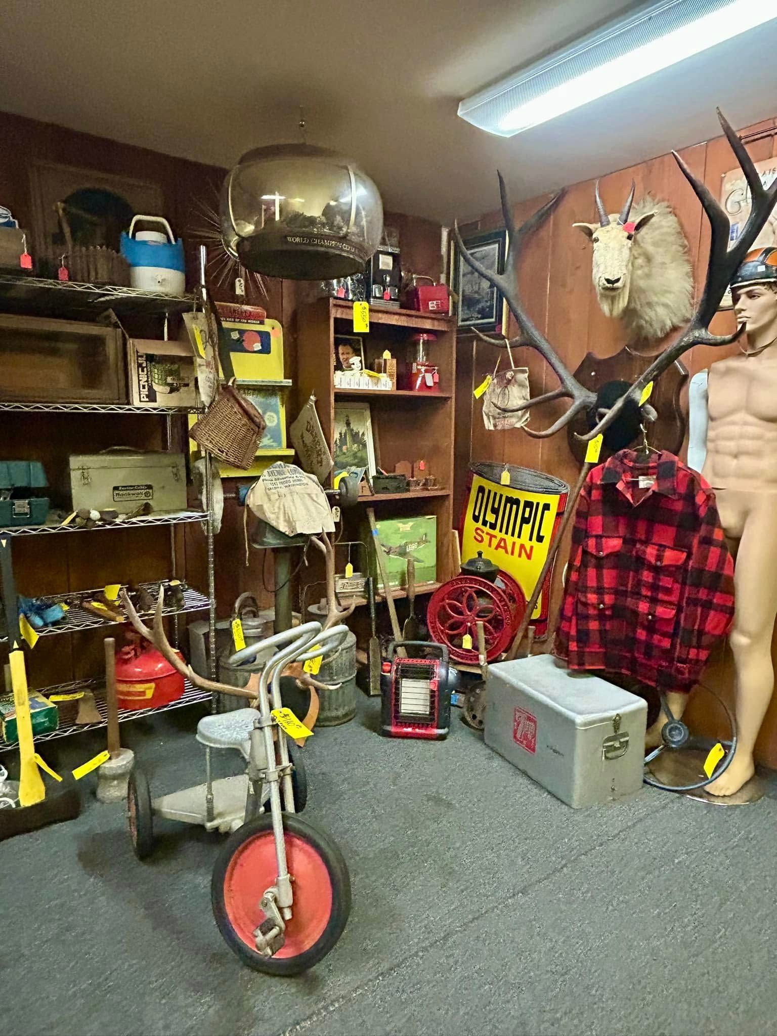 A cluttered antique store interior: shelves with items, a tricycle, mannequins, and various other vintage goods.