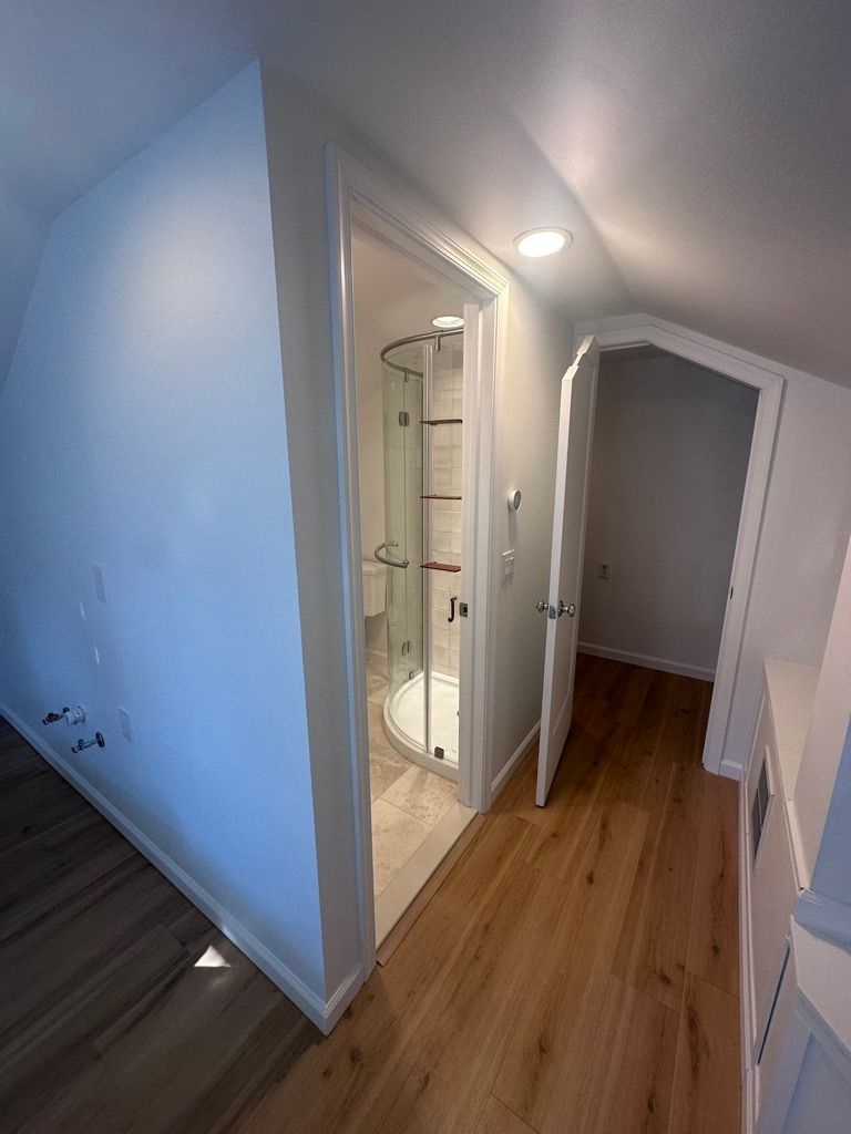 Hallway with wooden floor, leading to bathroom with glass shower and another doorway. Angled ceiling.