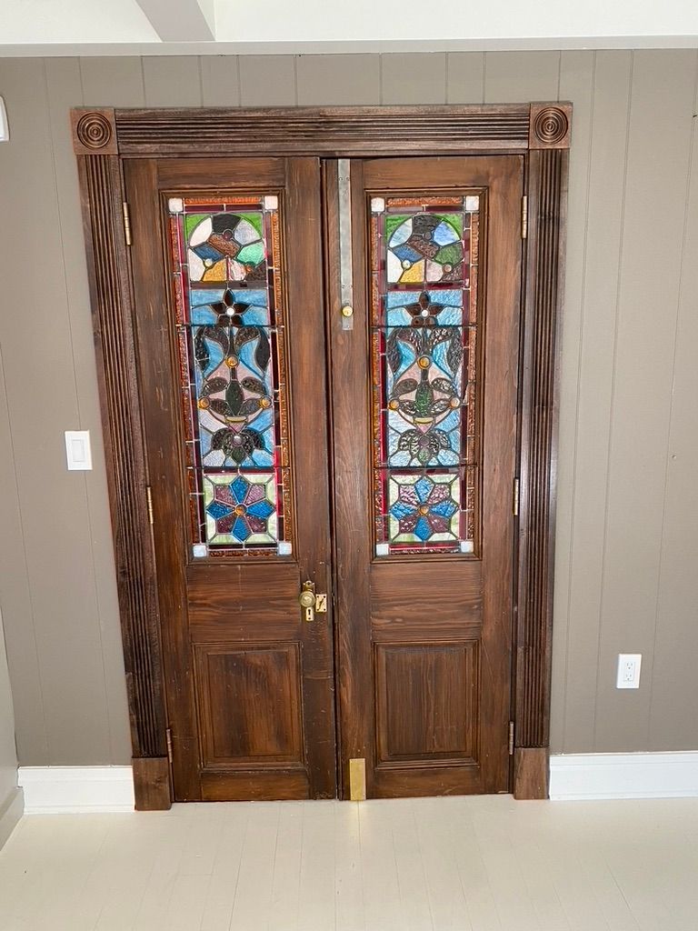 Wooden double doors with stained glass panels. The doors are brown and sit against a gray wall.