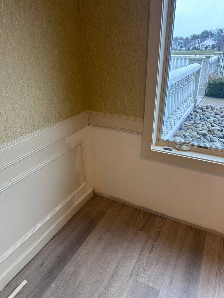 Corner of a room with wainscoting and wallpaper. A window looks out to a white balcony. Gray flooring.