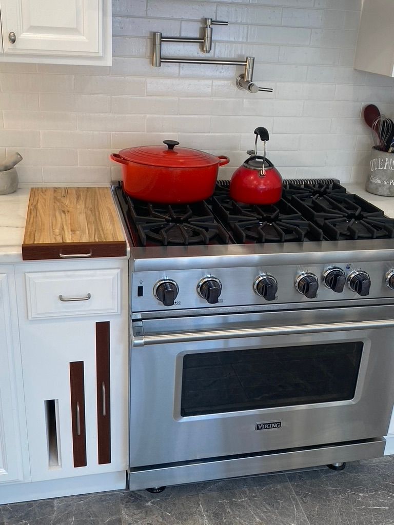 Stainless steel stove with red pot and kettle on stovetop, wooden cutting board, white cabinets.