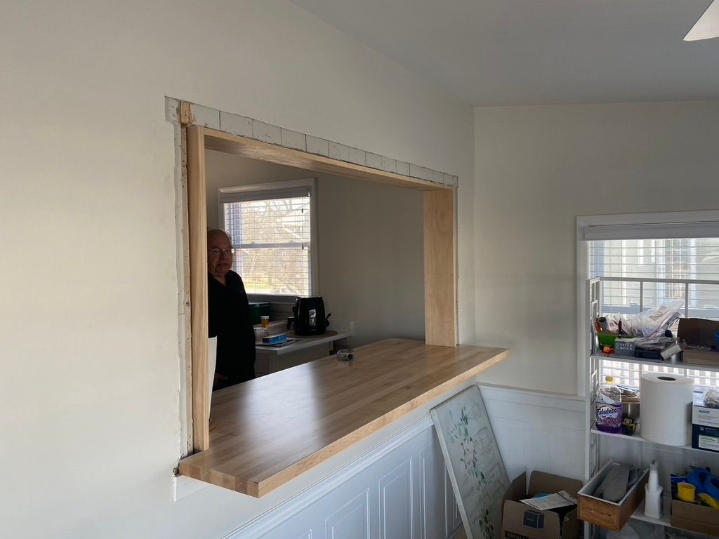 Open pass-through between rooms with wood counter. Man is visible in the adjacent room. White walls.